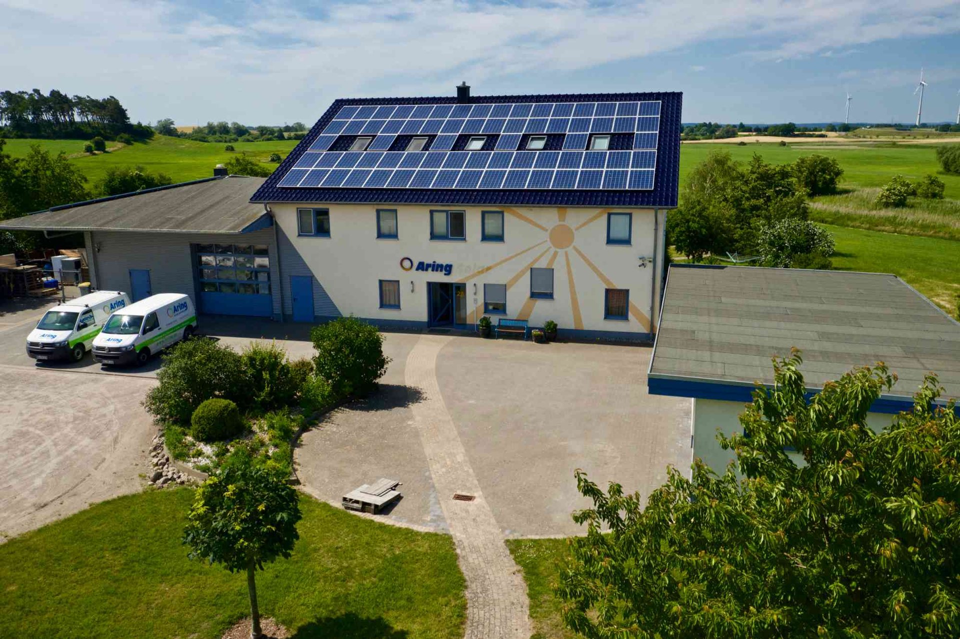 Firmengebäude mit Solarpaneelen auf dem Dach, zwei weißen Lieferwagen und einem gemalten Sonnenlogo.