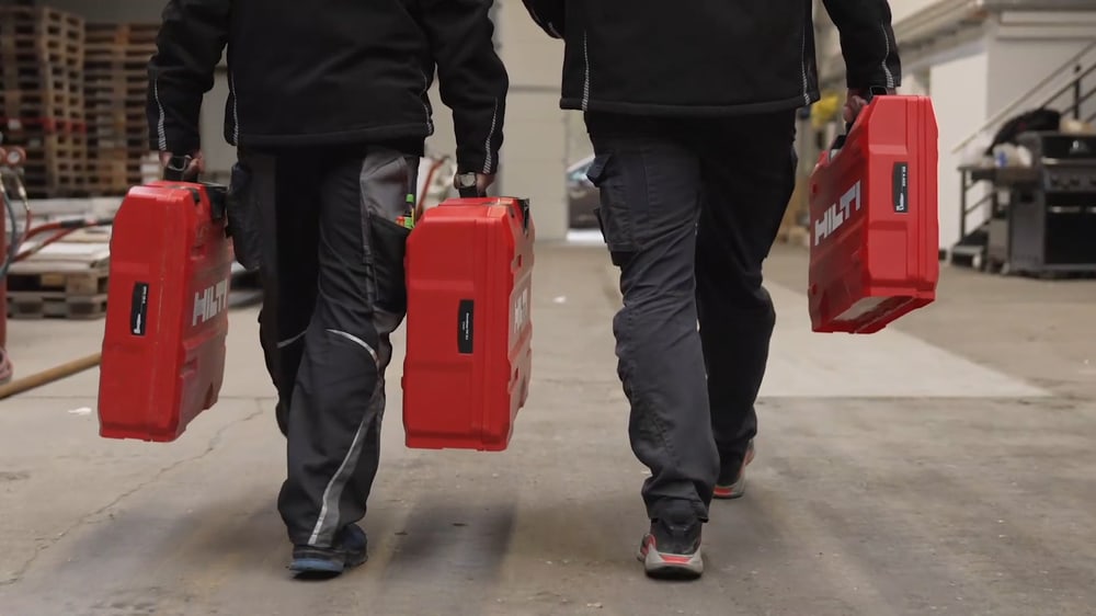 Zwei Arbeiter tragen rote Hilti-Werkzeugkoffer durch eine Lagerhalle.